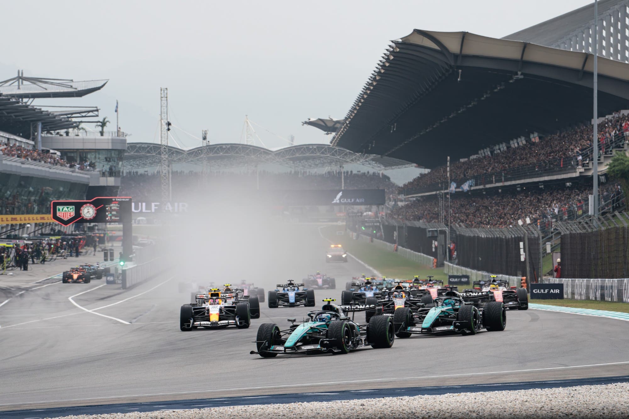 The field heads into Turn 1 in the wet at Sepang, in front of a packed main grandstand