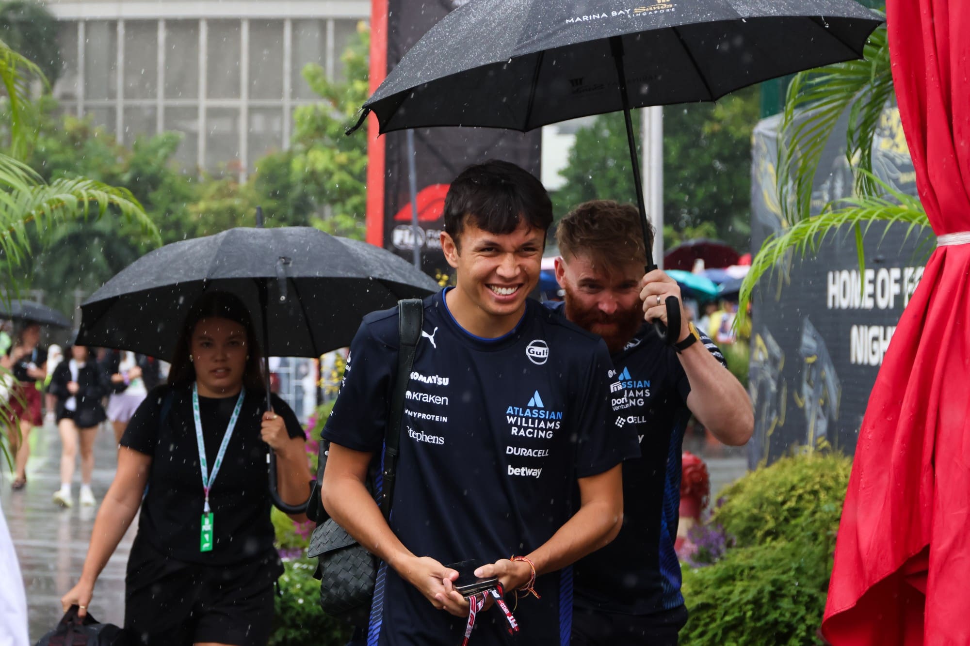 Alex Albon smiles as a Williams team member holds an umbrella over him in the rain outside the paddock