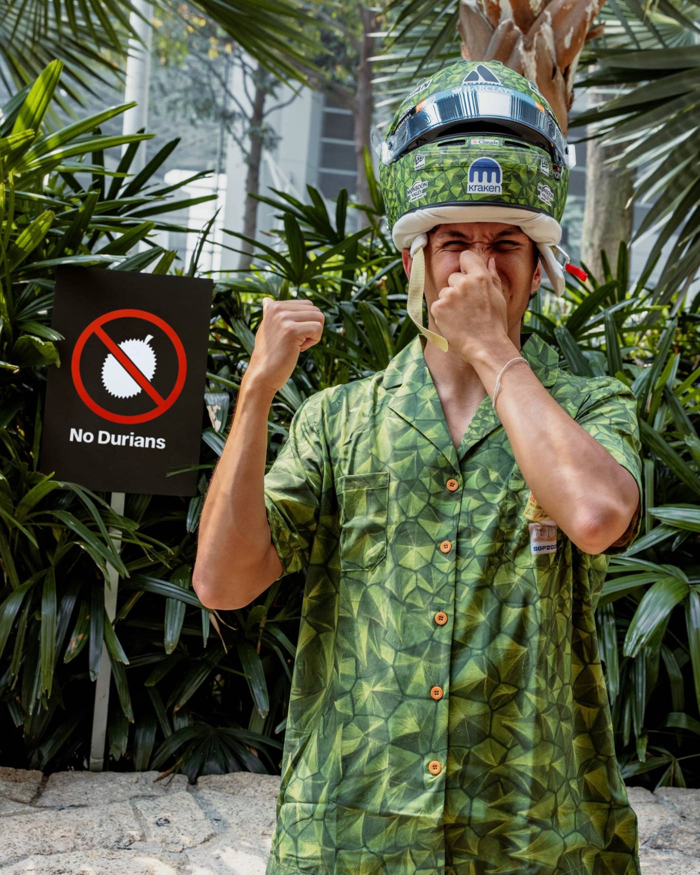 Alex Albon holds his nose in his durian helmet and shirt, next to a “No Durians” sign