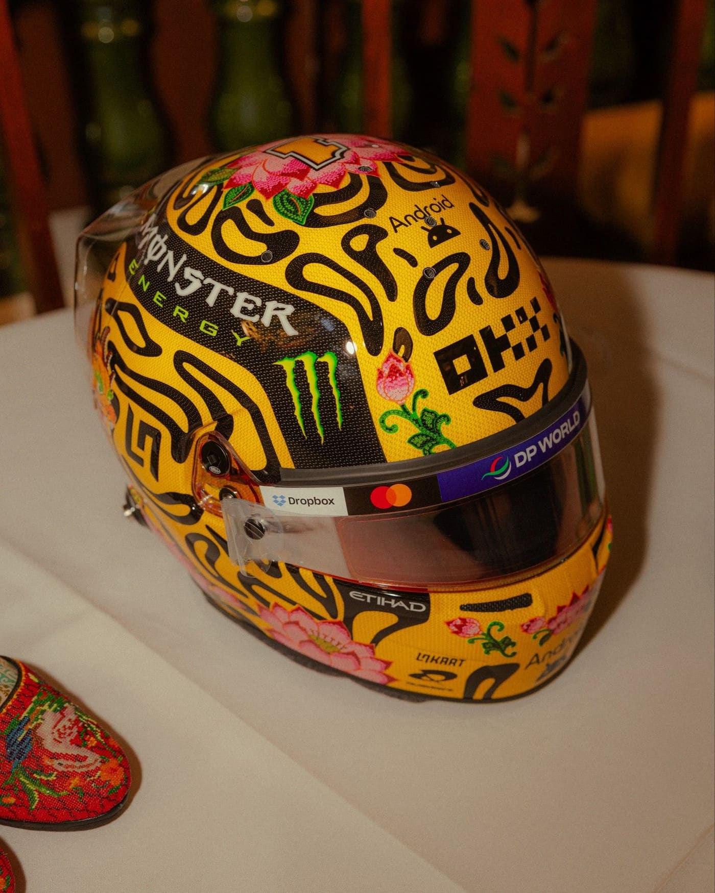 Lando Norris’s yellow helmet with black blobs and pink lotus flowers, next to a beaded Peranakan slipper