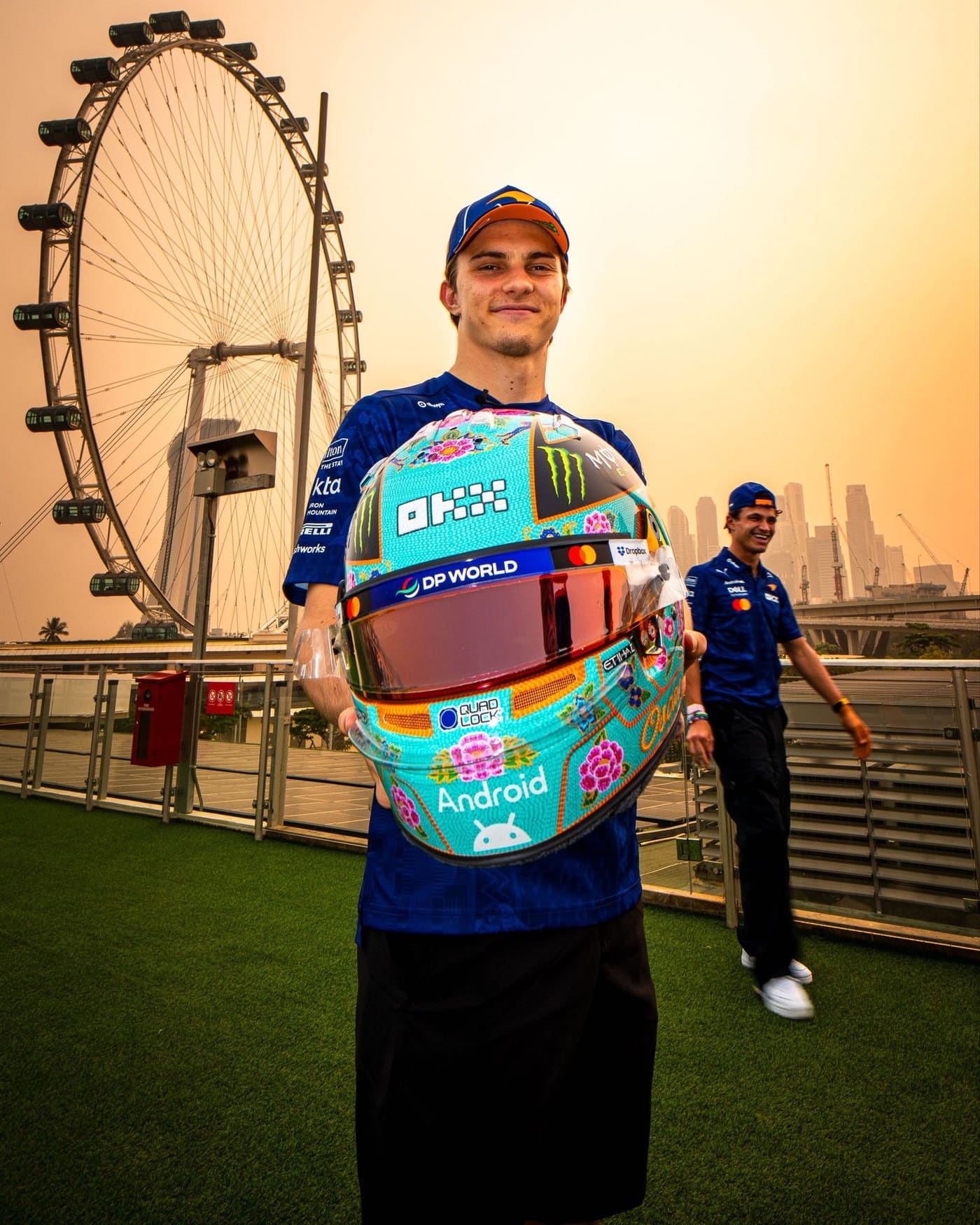 Oscar Piastri holds his beaded helmet in front of the Singapore Flyer, with Lando Norris behind him